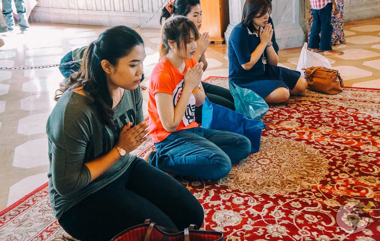 Grupo de mulheres ajoelhadas e com as mãos unidas em gesto de oração sobre tapete em área interna de templo.