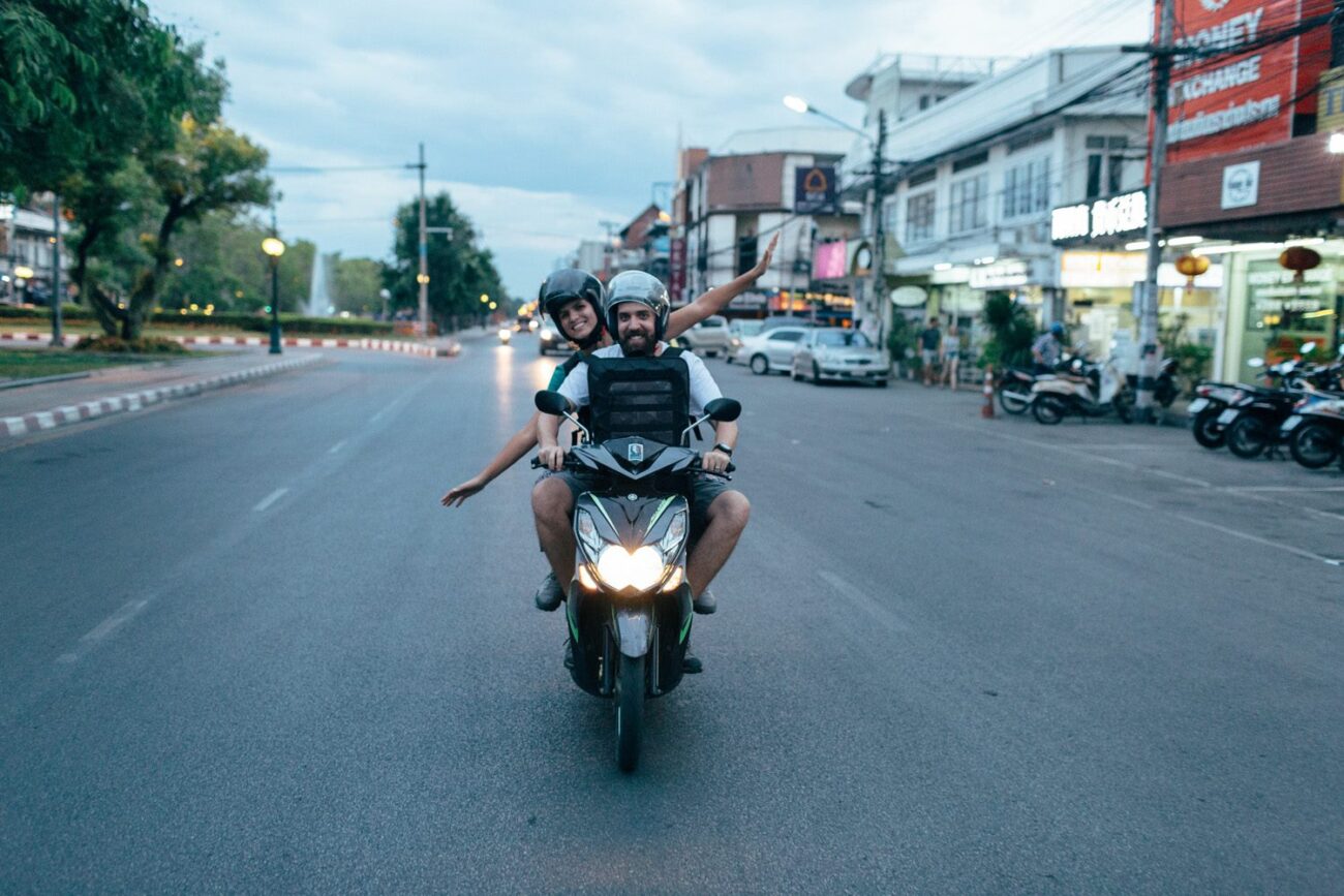Casal pilotando scooter em uma rua urbana da Tailândia no fim da tarde.