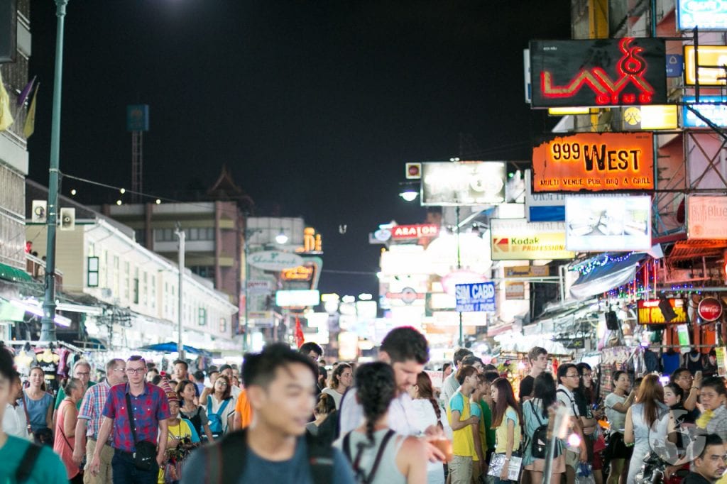 Multidão caminhando pela Khao San Road à noite, rodeada por letreiros luminosos e lojas abertas.