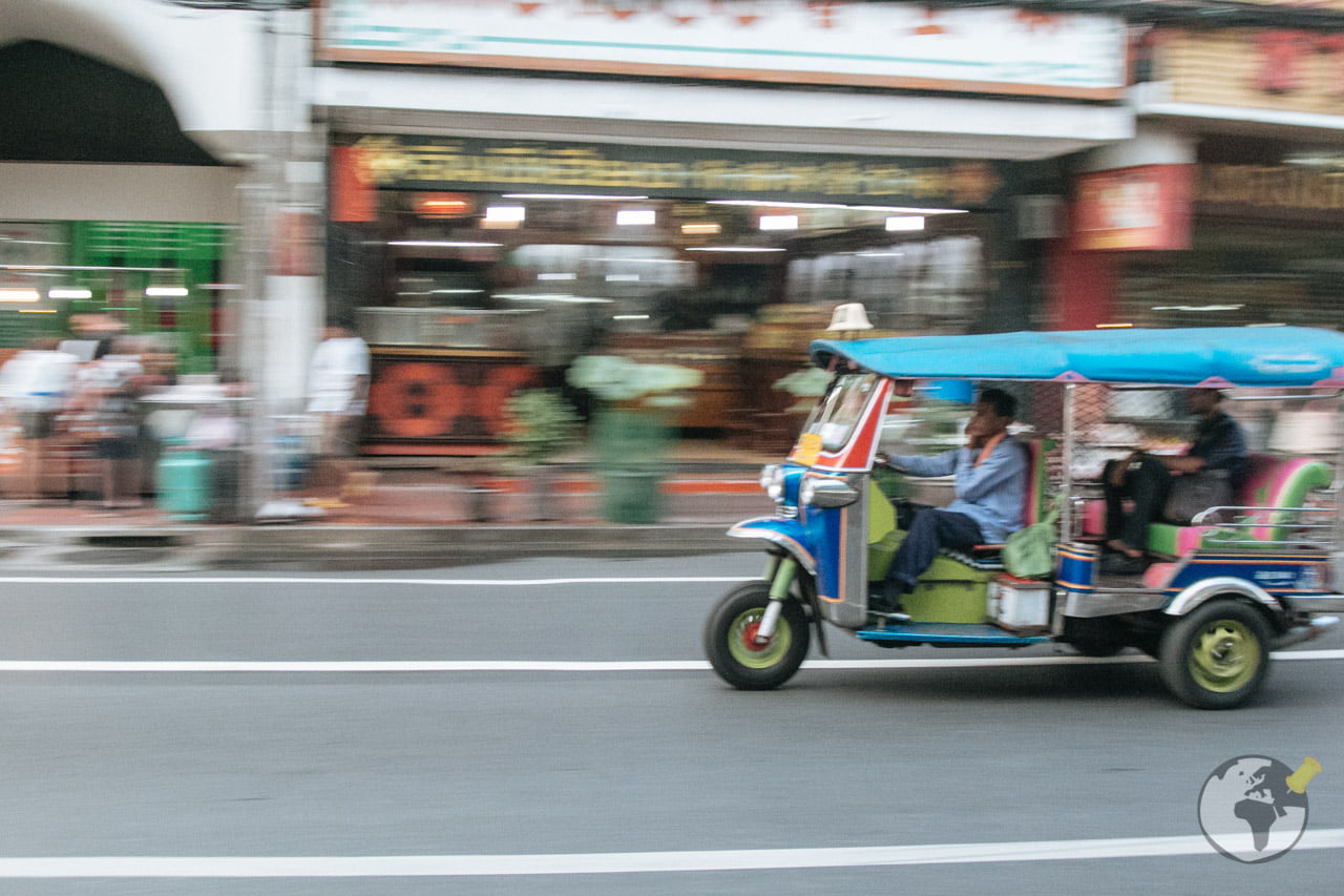 Tuk-tuk azul e verde circulando pelas ruas de Bangkok em frente a comércios locais.
