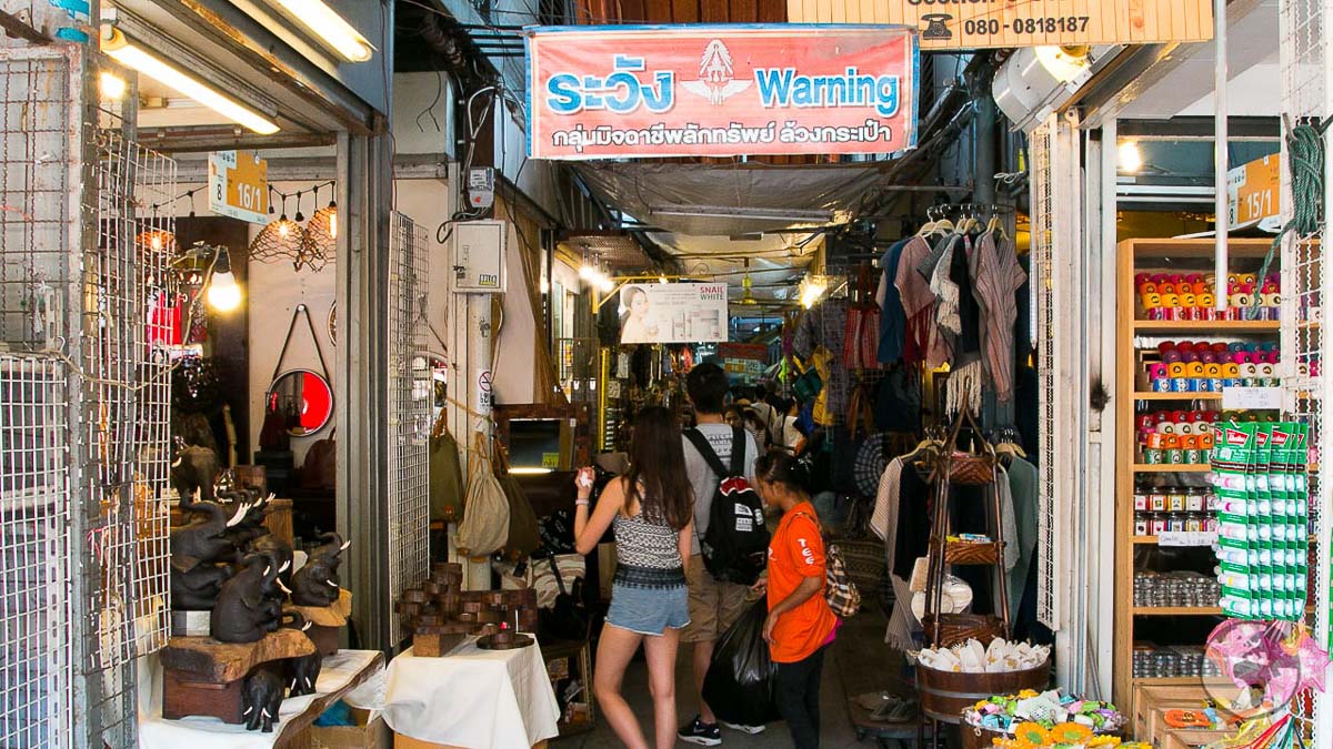 Corredor estreito do mercado de Chatuchak com turistas entre bancas de roupas, artesanato e produtos diversos.