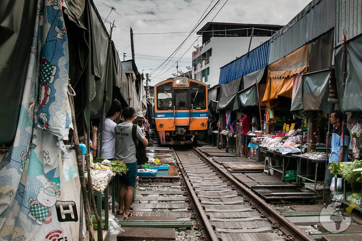 Trem laranja passando no meio do Mercado Maeklong, entre barracas com frutas, verduras e curiosos turistas.
