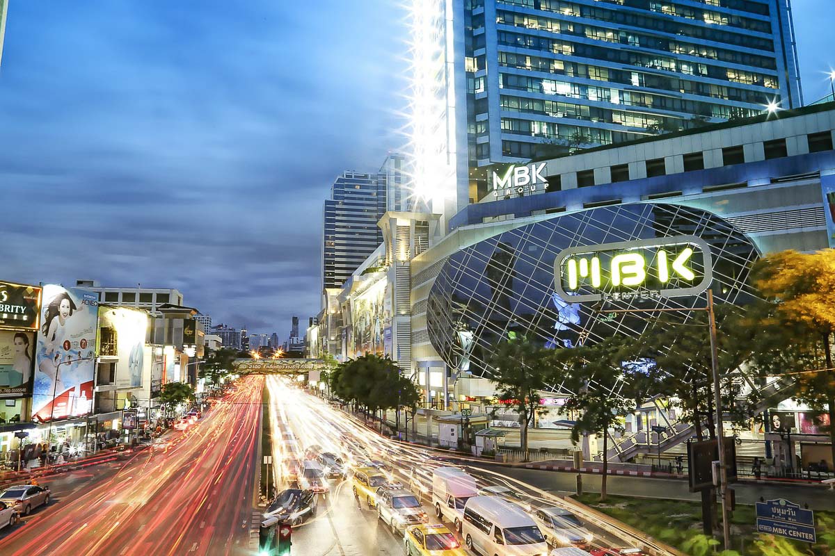 Fachada iluminada do MBK Center em Bangkok ao anoitecer, com trânsito intenso e rastros de luz dos carros.