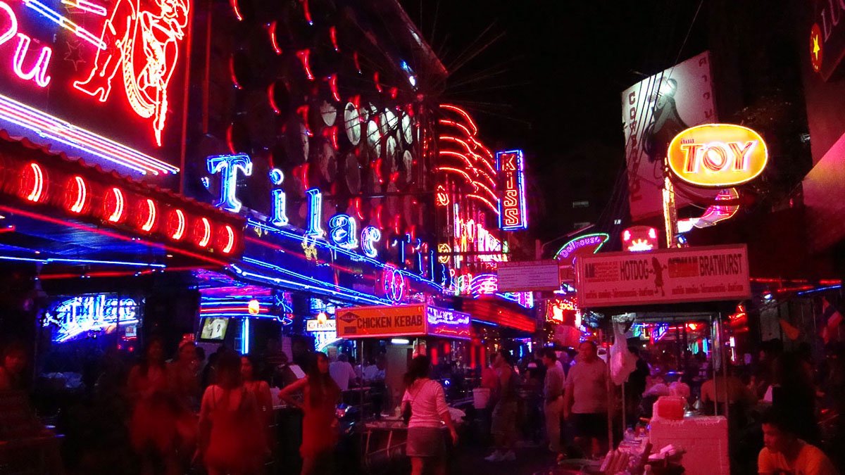 Rua movimentada de Bangkok à noite com letreiros de neon vermelhos e azuis e várias pessoas caminhando.