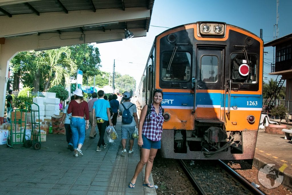 Dri sorrindo ao lado de trem laranja e azul parado na estação de Kanchanaburi.
