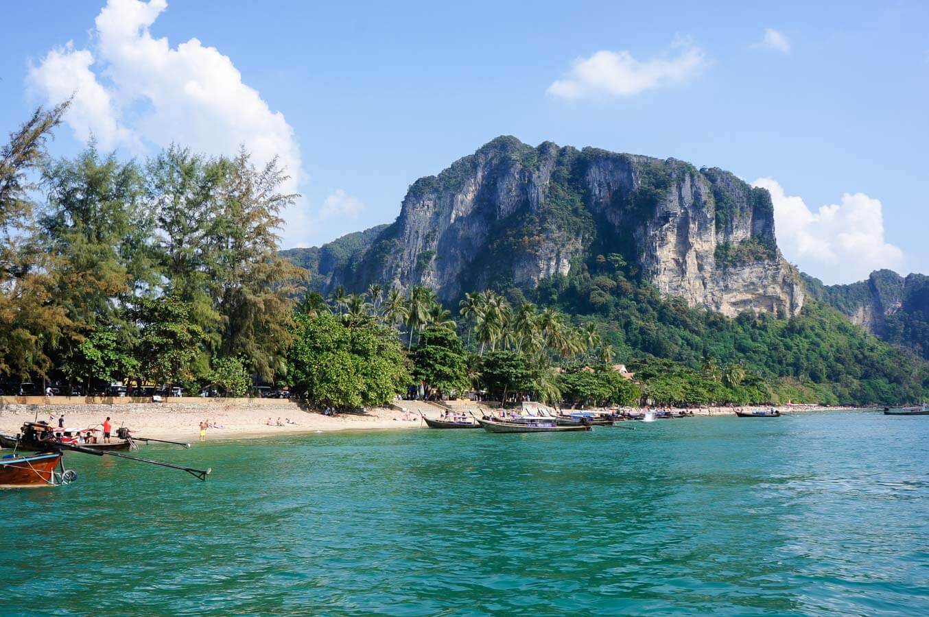 Praia com barcos longtail, vegetação tropical e penhascos ao fundo em Ao Nang, Krabi.