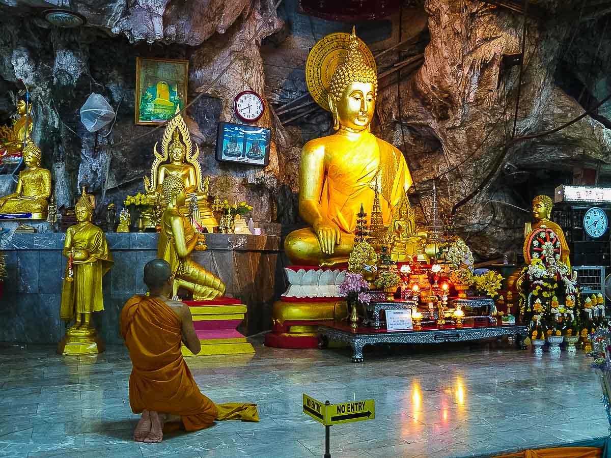 Monge ajoelhado diante de estátuas douradas de Buda em templo dentro de caverna.