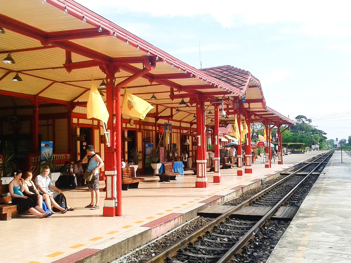 Estação ferroviária de Hua Hin com estrutura em vermelho e amarelo, onde alguns viajantes aguardam sentados na plataforma.