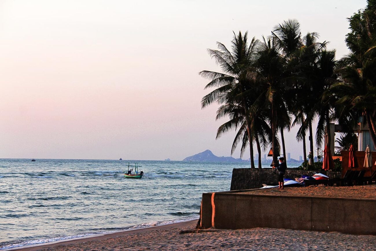 Barco pesqueiro navega próximo à orla da Praia de Hua Hin, com palmeiras e céu rosado ao entardecer.