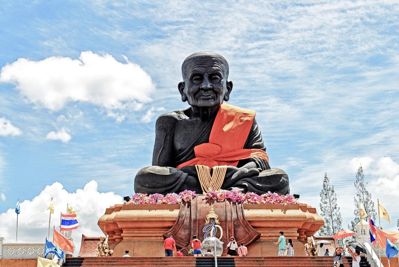 Estátua gigante do monge Luang Phor Thuad no Wat Huay Mongkol, com visitantes na base e céu azul ao fundo.