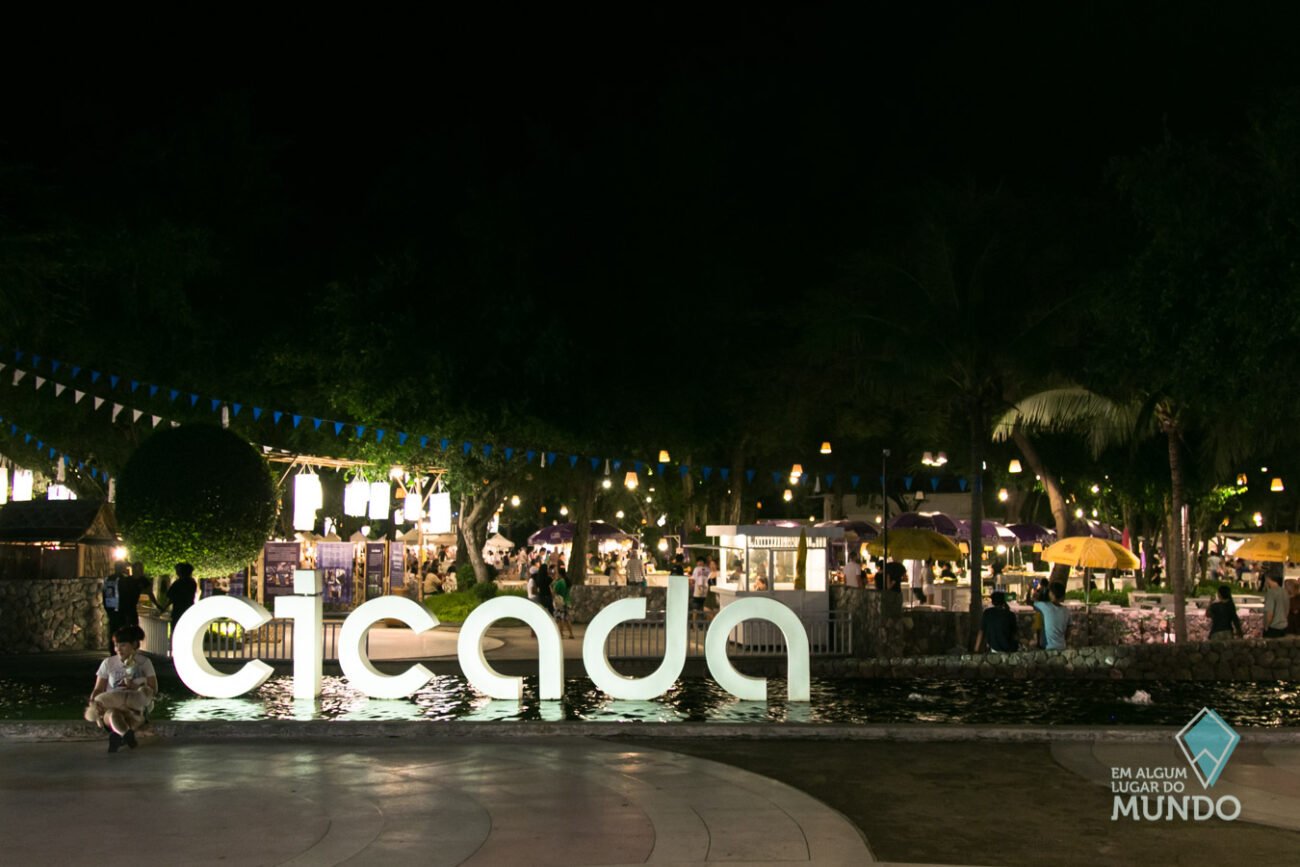 Letreiro iluminado com a palavra “Cicada” em frente à entrada do mercado noturno, com tendas, bandeirinhas e visitantes ao fundo.