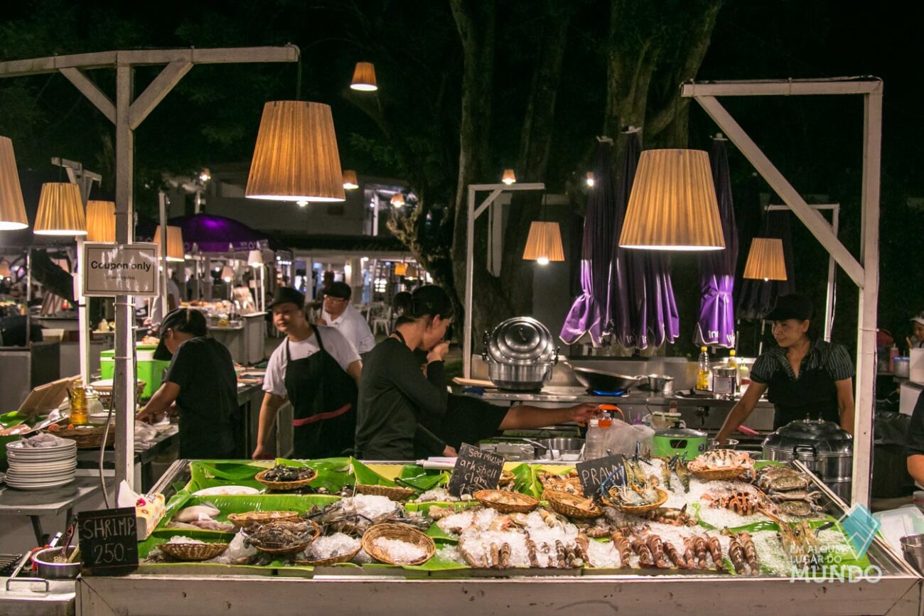 Bancada de frutos do mar frescos expostos com gelo no Hua Hin Night Market, com vendedores ao fundo preparando pratos.