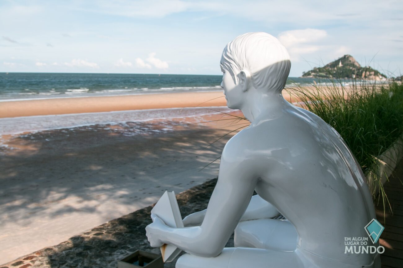 Escultura branca de uma pessoa sentada com um livro nas mãos observa o mar tranquilo da Praia de Hua Hin.