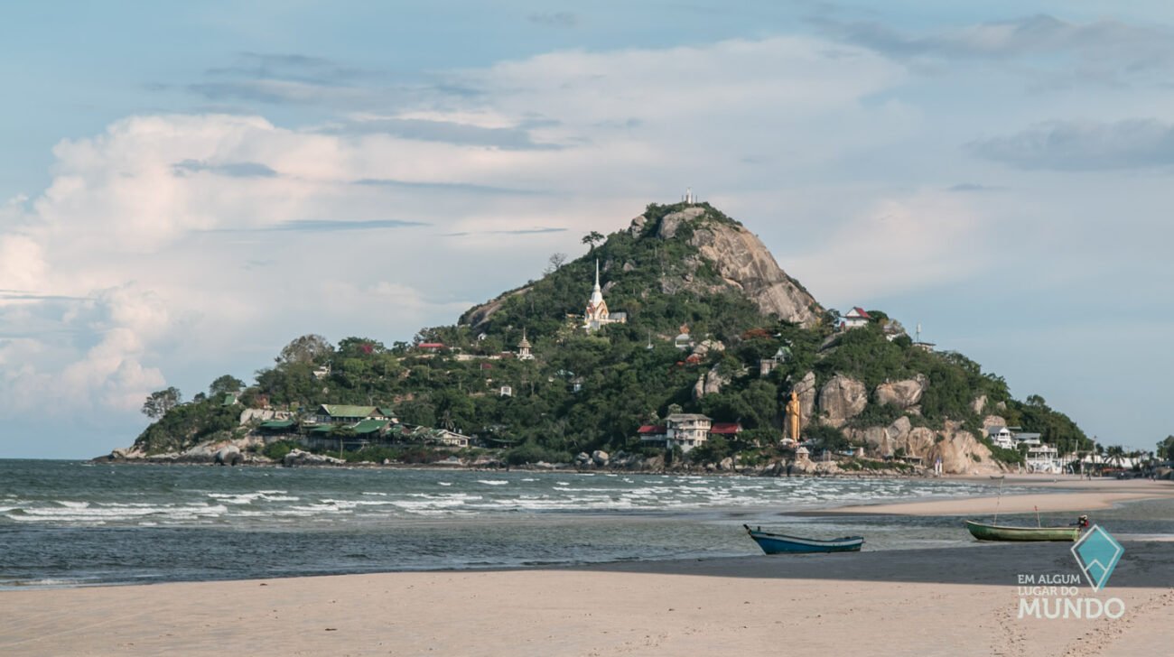 Vista da colina de Khao Takiab a partir da praia, com templos e construções espalhadas pela encosta e uma grande estátua dourada ao pé do morro.