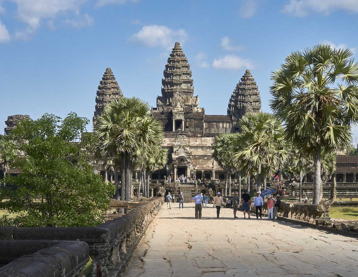 Entrada principal de Angkor Wat com turistas caminhando pela passarela de pedra, rodeada por palmeiras.