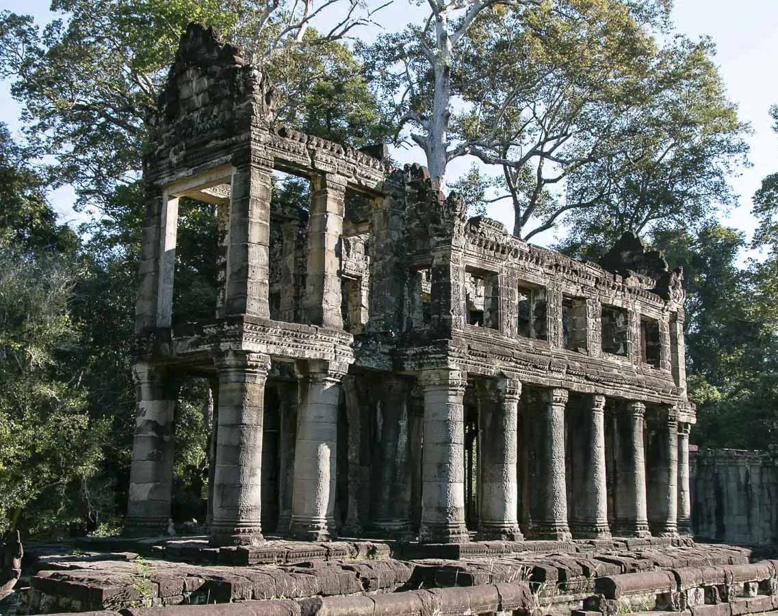 Ruínas do templo Preah Khan com colunas de pedra e árvores altas ao fundo.