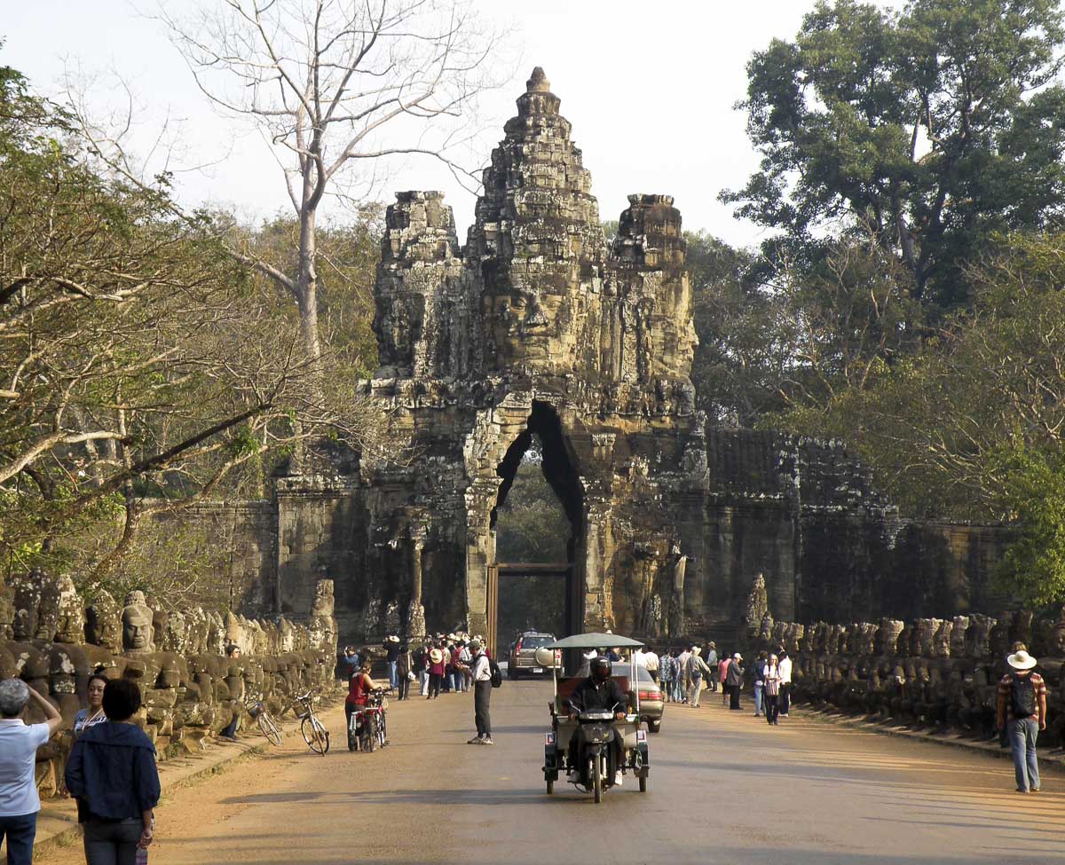 Entrada monumental de Angkor Thom com rostos esculpidos e turistas passando em tuk-tuks.