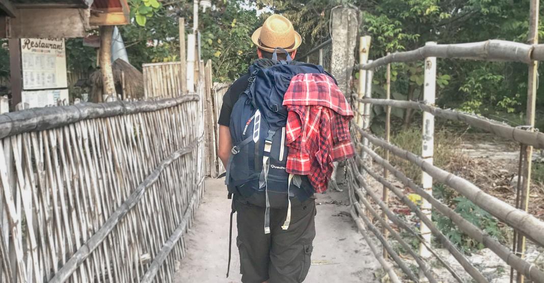 Caio caminha em direção a uma vila com mochila grande nas costas e uma camisa amarrada.