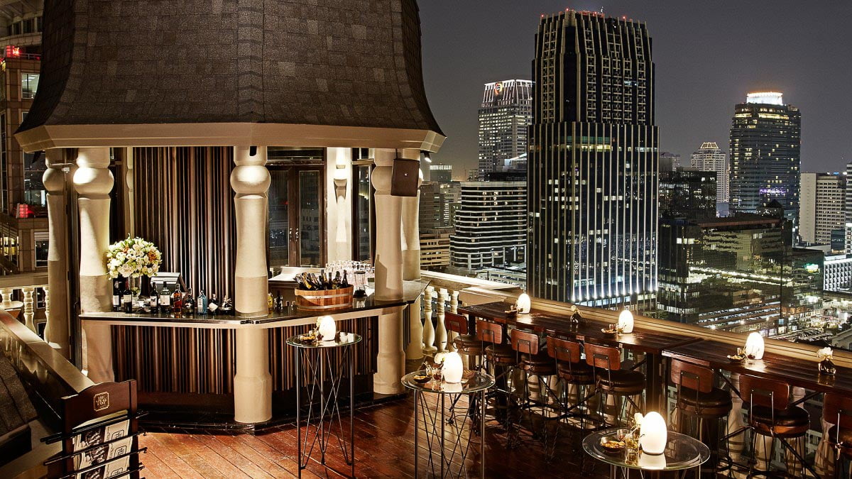 Speakeasy Rooftop Bar com ambiente elegante, bar em madeira e vista panorâmica de Bangkok à noite.