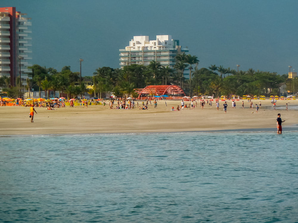 Uma parte do mar e ao fundo a areia e banhistas na Praia da Enseada
