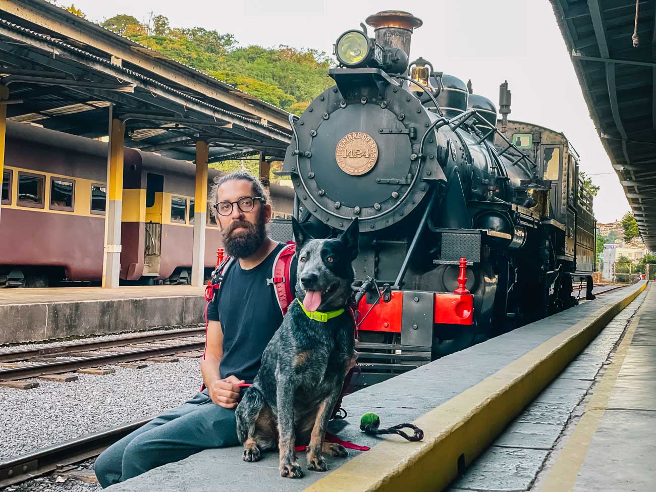 Homem sentado com um cachorro na Estação Ferroviária de São Lourenço e ao fundo a locomotiva