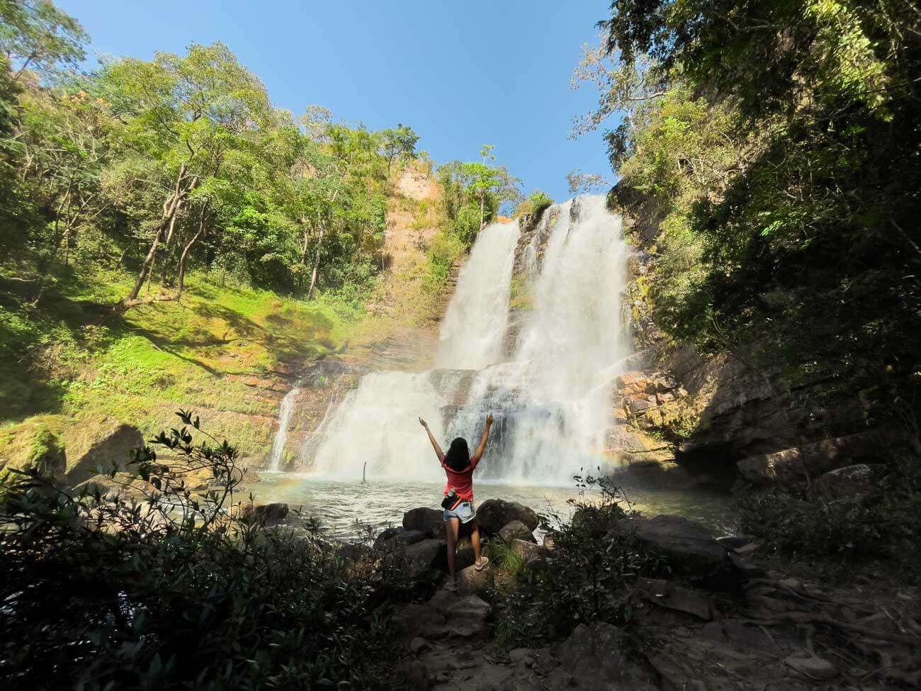 Mulher em pé de braços abertos para cima em frente à queda da Cachoeira Véu da Noiva na Chapada Indaía