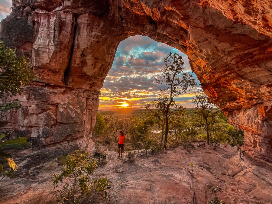 Mulher com um cachorro na abertura do Arco do Sol, e ao fundo o pôr do sol e a vegetação do cerrado
