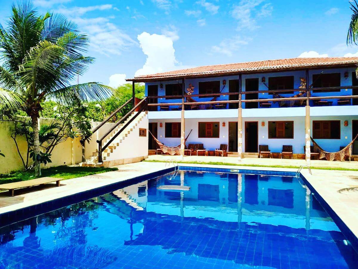 Piscina ao ar livre com alguns coqueiros na lateral e pousada ao fundo