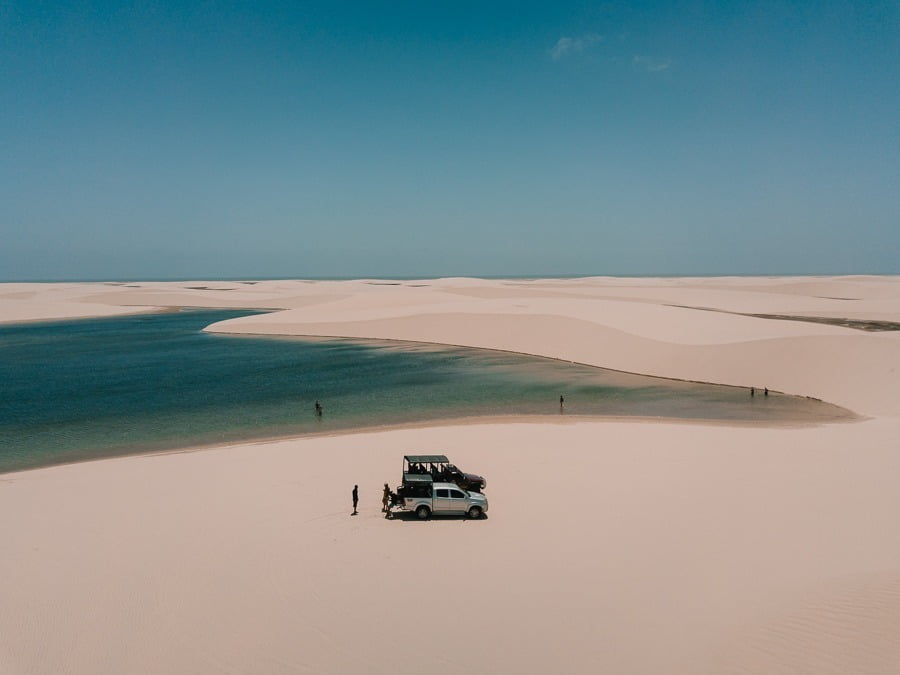 Vista de cia das dunas dos Lençóis Maranhenses com lagoas de águas azuis. Há dois carros parados e pessoas próximas deles.