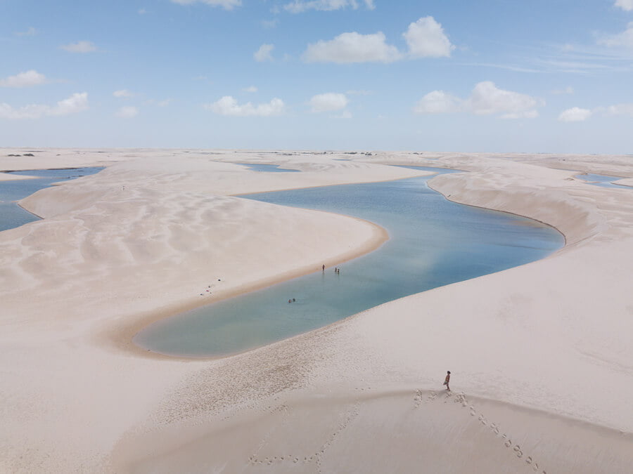 Lagoa do S nos Lençóis Maranhenses vista de cima com águas em tons de azul claro e uma mulher caminhando no topo da duna ao redor