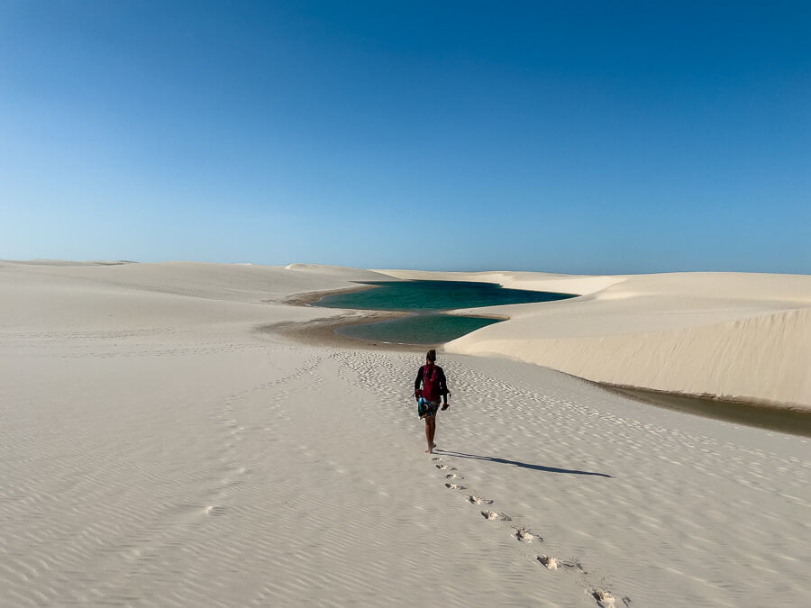 Um homem caminhando em dunas de areia branca nos Lençóis Maranhenses , ao fundo uma lagoa.