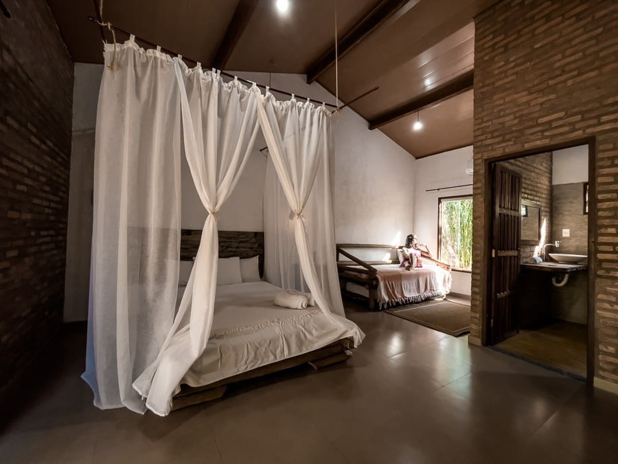 Cama de casal com mosquiteiro e decoração rústica. Ao fundo uma mulher sentada em um sofá olhando pela janela da pousada Rancho das Estrelas
