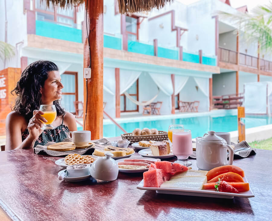 Mulher sentada em uma mesa repleta de itens de café da manhã em frente a piscina da Pousada Village Sonhos