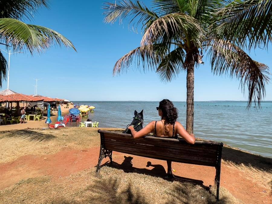 Mulher e cachorro sentados em banco de madeira em frente à Praia dos Arnos em Palmas