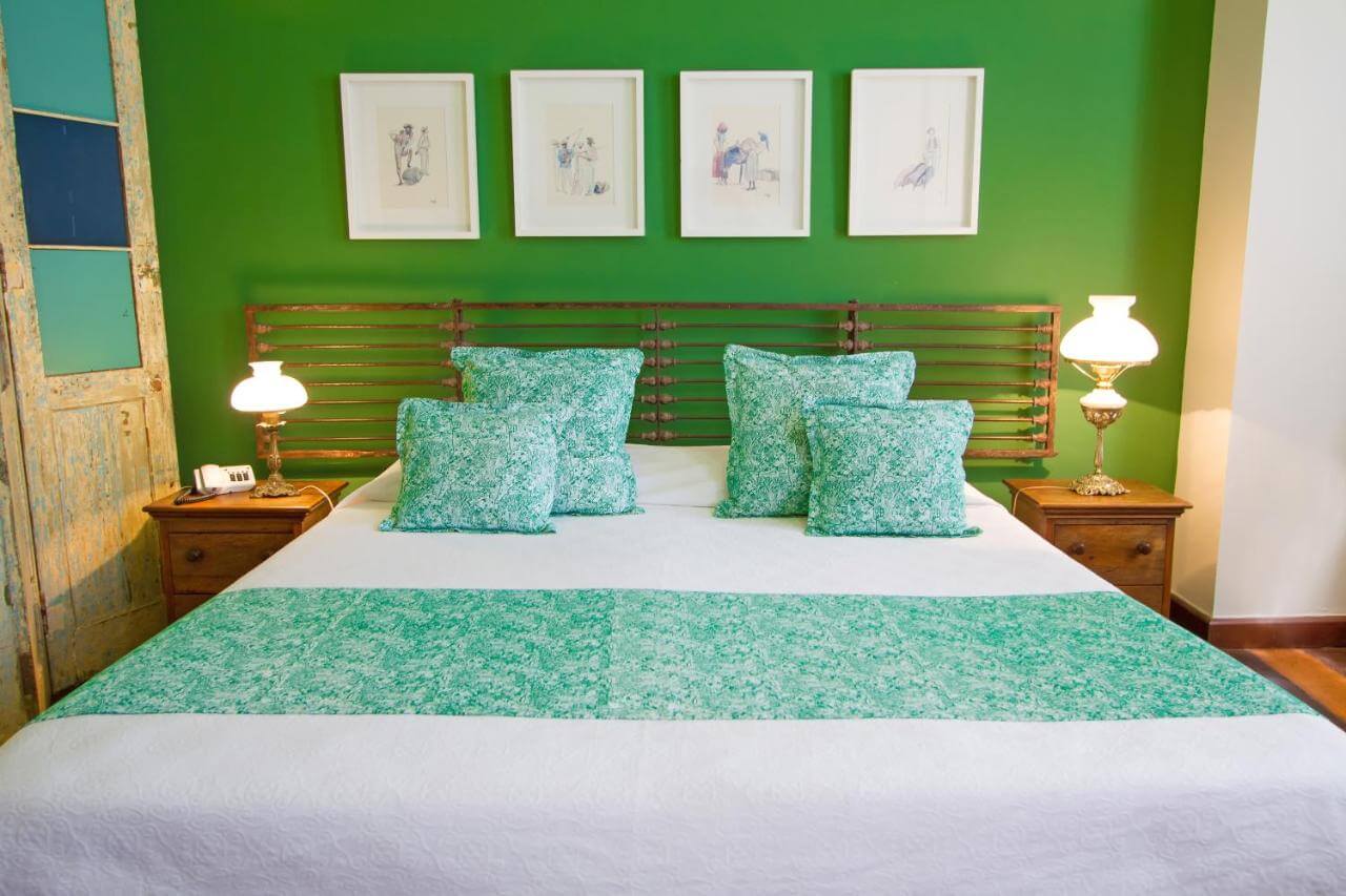 Cama de casal com cobre leito em detalhes verdes e parede ao fundo verde em pousada em Salvador