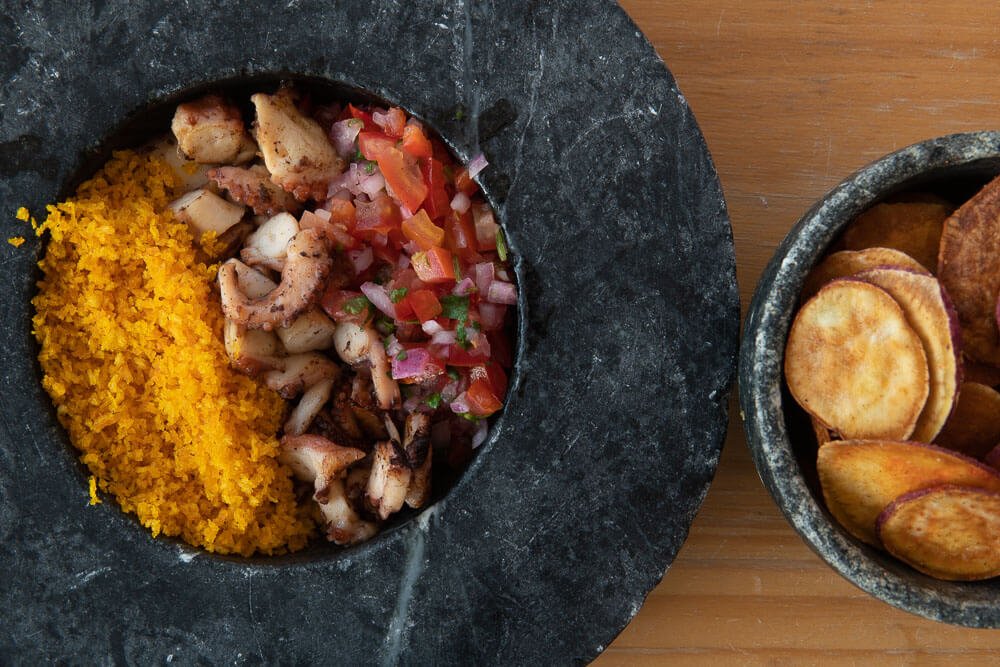 Prato com pedaços de polvo grelhado, molho vinagrete e farova de farinha panko. Ao lado um bowl de chips de batata doce.