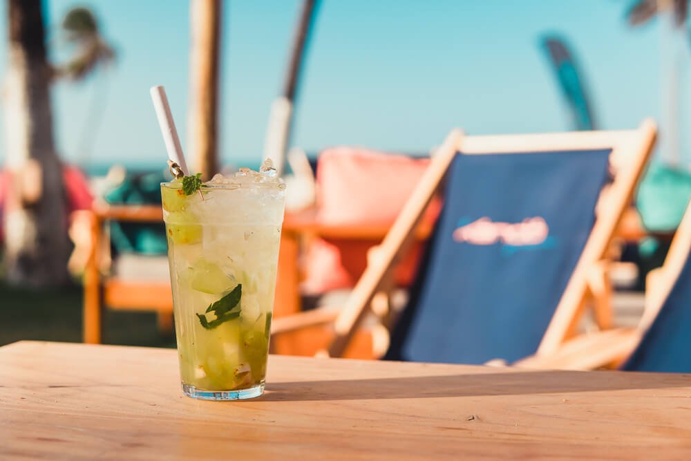 Copo de caipirinha sobre mesa de madeira com cadeiras espreguiçadeiras ao fundo no sol.