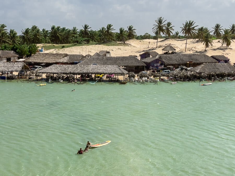 Lago Grande em Camocim com águas claras e cristalinas. Ao fundo redes e restaurantes rústicos próximos à margem e dunas