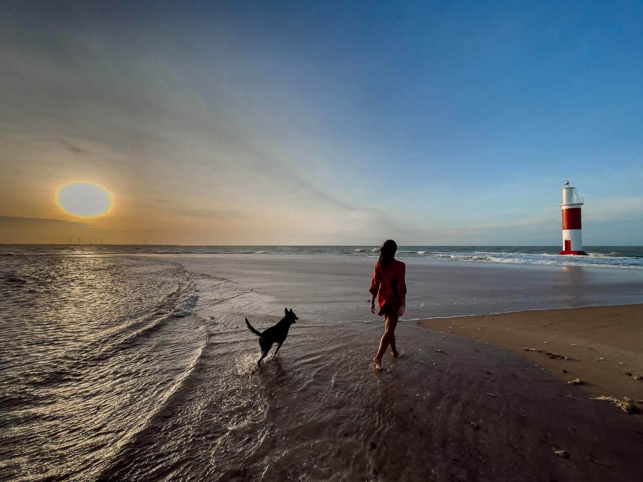 Uma mulher e um cachorro caminhando em direção ao farol nas areias da Praia do Farol