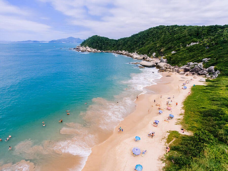 Vista de cima da Praia da Tainha em Bombinhas com areia clara com algumas barracas e um morro com costão e vegetação ao fundo