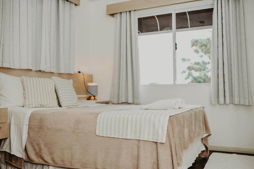 Cama com manta rosé e ao fundo janela de vidro em pousada em Bombinhas
