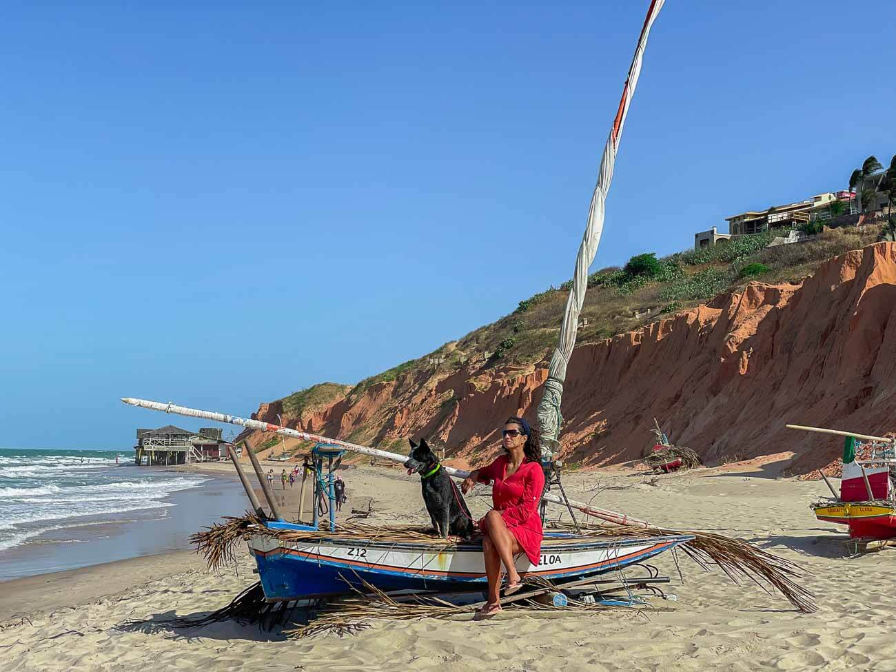Mulher e cachorro sentados em barco parado na areia da Praia de Canoa Quebrada