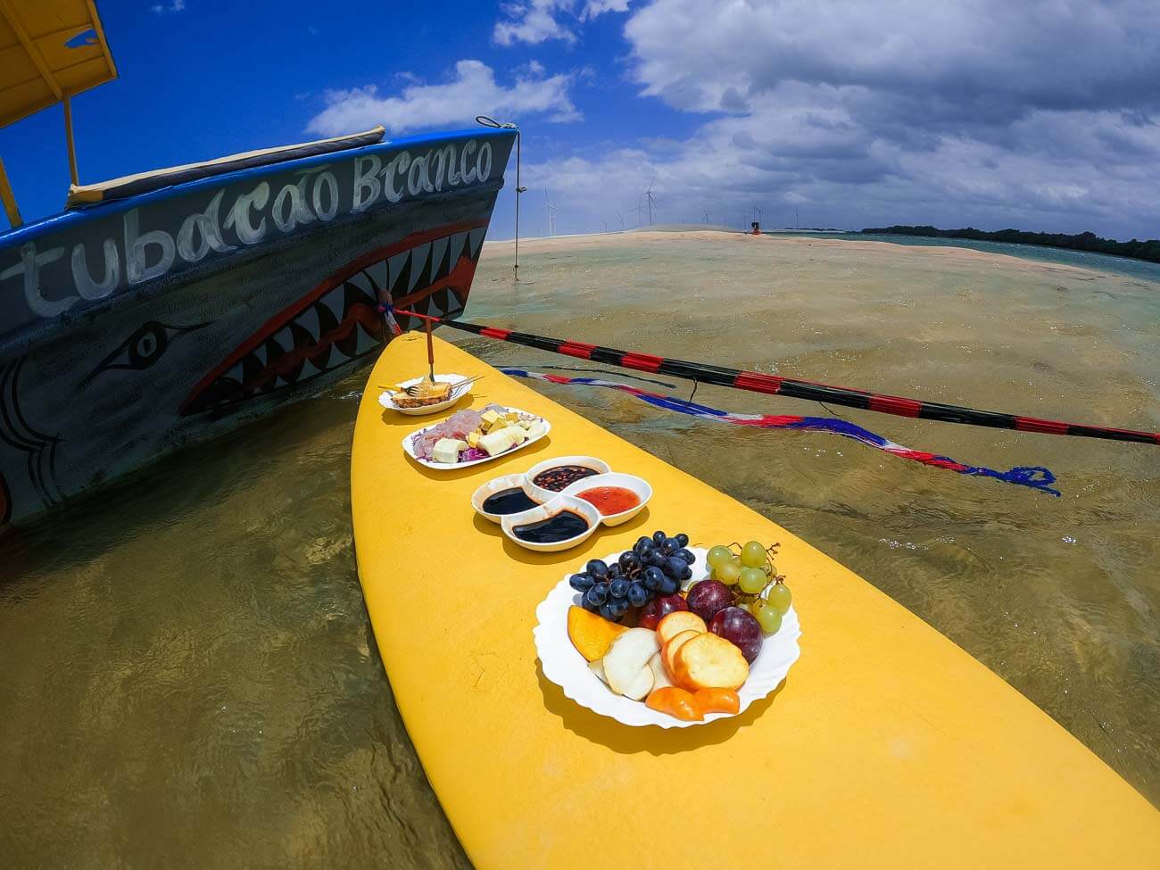 Uma prancha amarela na água com frutas, geleias e frios em cima durante passeio de barco em Galinhos