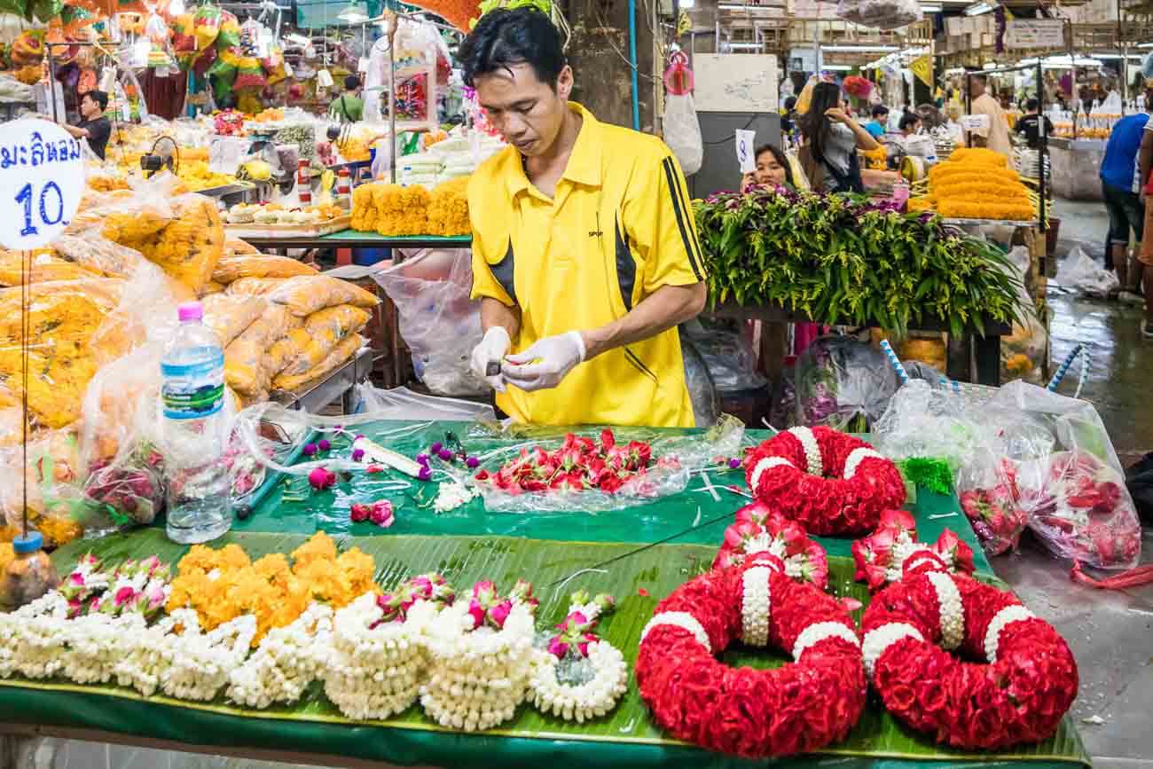 Homem preparando colares de flores vermelhas e brancas em uma barraca do mercado de flores de Bangkok.
