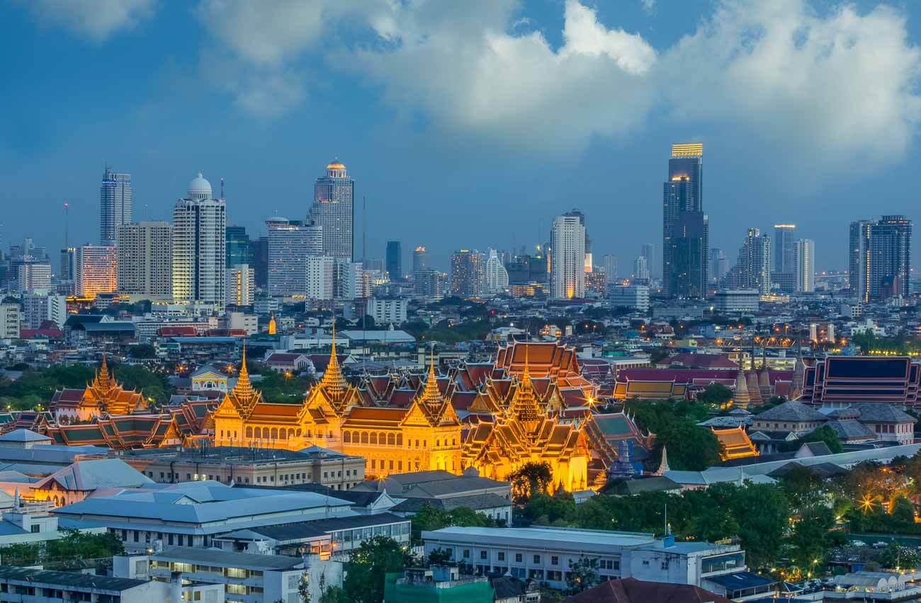 Grand Palace iluminado ao anoitecer com os arranha-céus de Bangkok ao fundo.