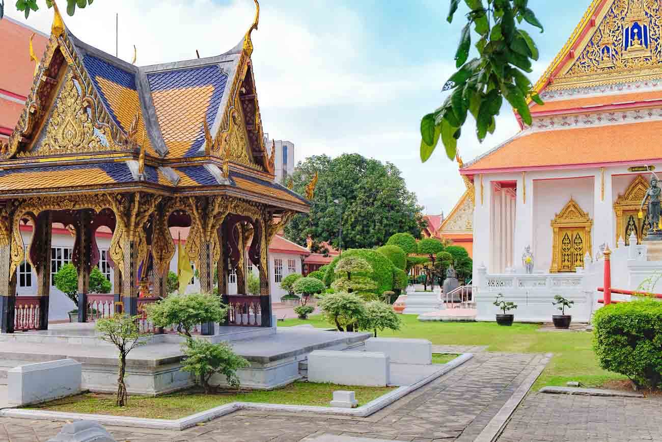Pavilhão ornamentado no jardim do Museu Nacional de Bangkok com templo ao fundo.