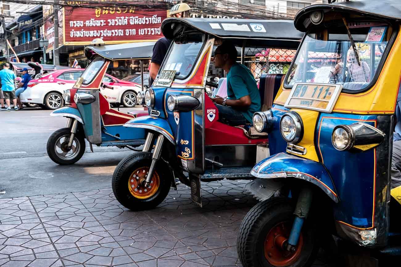 Vários tuk-tuks coloridos estacionados na rua com motoristas esperando passageiros em Bangkok.