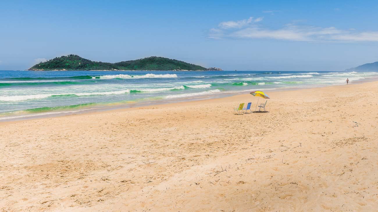 Praia do Campeche com larga faixa de areia e um guarda roupa com duas cadeiras de praia.