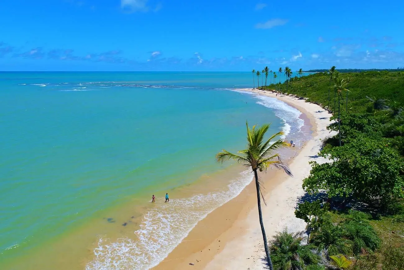 Vista de uma parte da Praia de Cumuruxatiba com a orla repleta de vegetação litorânea e coqueiros.