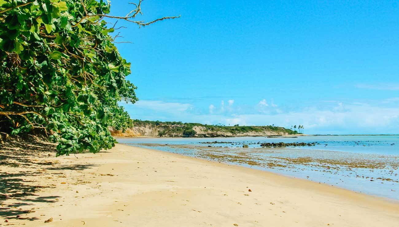 Praia do Peixe Grande em cm areia vazia e o mar calmo. Ao fundo, dunas com vegetação.